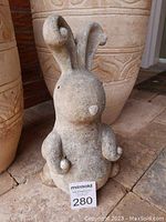 Front view of faux stone resin bunny statue with curled ears, showing rough textured surface sitting on brick tiles with large pottery in background.