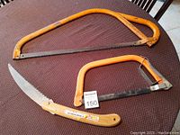 Three saws displayed on a brown textured surface: a large 29 inch yellow bow saw, a smaller 16 inch yellow bow saw, and a folding pruning hand saw with wooden handle and serrated blade.