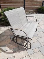Side angled view showing brown metal frame and grey fabric seating of the swing glider loveseat on patio pavers.