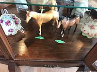 Overview of two Beswick horse figurines placed between two floral porcelain footed bowls on display stands inside a wooden cabinet.