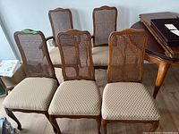 Photo showing all five wooden chairs, three side chairs and two captain chairs, arranged together with visible caned backs and beige upholstered seats.
