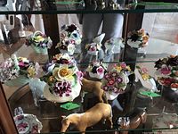 Wide shot of glass shelf displaying multiple porcelain floral baskets and decorative plates along with some horse figurines underneath