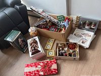 Photo showing assorted Christmas decorations including a box of wrap and tins, framed Santa pictures, ornaments, figurines, and a jar of ribbons.