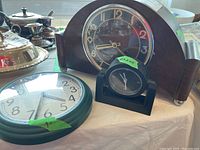 Photo showing the three clocks: wooden mantle clock, black desk clock, green framed wall clock, placed on a table.