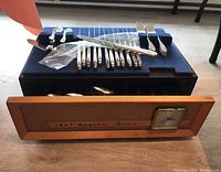 Front view of the wood case open with flatware displayed on blue velvet tray and the built-in square Mauthe alarm clock visible on the front panel.