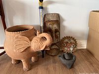 Front left side view of clay elephant planter with wooden mask, clay sun mask, and stone mortar and pestle in background.