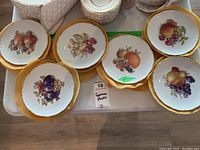 Twelve plates displayed with different fruit designs on them with gold scalloped rims.