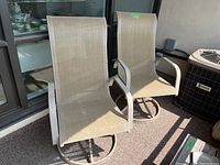 Photo showing two metal swivel patio chairs with beige mesh seats on a balcony next to a window and air conditioning unit.