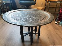 Metal round tray table assembled, showing ornate surface and wooden folding legs.