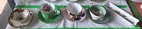 Four tea cups and saucers and one long narrow dish arranged side by side on a white cloth with green tape.