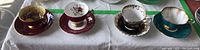 Long shot showing four tea cup and saucer sets arranged in a row: Aynsley, Adderley, and two Royal Albert.