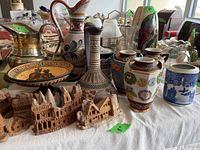 Overall view of ceramics, glass vases, metal and ceramic pitchers, and wood miniature village models on table.