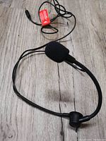 Black headset microphone with foam windscreen, adjustable boom arm, cable with 3.5mm plug lying on light wood floor.