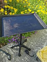 Full view of black rectangular tray table on 5-legged base, showing height adjustment and mobility with caster wheels. Tray surface has faded areas and scratches.