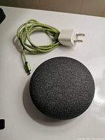 Google Nest Mini smart speaker in charcoal fabric, with attached braided USB charging cable and white power adapter plug, placed on white surface.