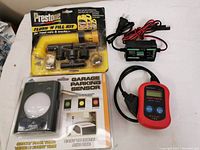 Photo of all four items: OBD2 scan tool, parking sensor in package, antifreeze flush kit in sealed blister pack, and trickle charger with cords