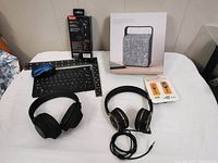 Photo showing all items including wireless speaker in box, two headsets, keyboard, mouse, and batteries on white fabric surface.