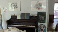 Four artworks shown above a piano: two are framed and hanging on the wall; two rest on the piano, including a canvas and an art board painting.
