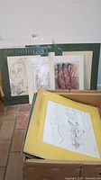 Photo showing multiple signed artworks with mats leaning against a wall, and a box containing additional artworks in yellow mats.