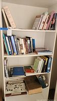 Photo of bookshelf showing three shelves filled with various books, including large reference books and smaller paperbacks covering history, geography, fiction, and birds.