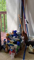 Shows multiple spray bottles and liquid cleaning products grouped with brooms and mop handles visible at the back
