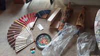 Full view of all items: five folded fans with different floral patterns, two decorative floral umbrellas, a Russian decorative plate, and a pin on carpeted floor.