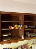 Wood cabinet shelves with stacked plates, bowls, and boxes of food wraps including plastic wrap, foil, and parchment paper.
