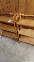 Pair of wooden folding and stackable bookshelves placed adjacent to each other on carpeted floor with wood-paneled wall background. Shows overall design and size.