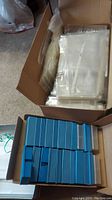 Open cardboard box filled with blue plastic slide storage boxes stacked neatly and another box containing numerous clear plastic slide protector sheets with multiple pockets per page.