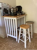 Photo of counter height table with natural wood top and white slatted sides with two stools beside it, showing condition of both stools and table