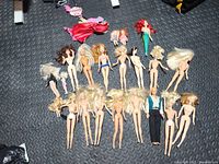 Top-down photo showing entire lot of 17 dolls, mostly nude Barbies with one Ken doll and one mermaid doll, arranged on black textured surface