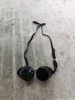 Full view of vintage welding goggles laid flat on a rough surface, showing dark lenses and adjustable strap.