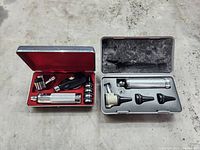 Two otoscope kits in metal cases with foam inserts. One has black and silver parts with several detachable pieces; the other has a white head and three black ear specula