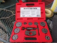 Photo showing an open red plastic case containing a disc brake caliper tool set including a screw-type central caliper piston tool and nine circular adaptor plates.