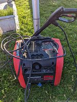 Husky red and black electric power washer with hose and spray gun resting on grass. Viewed from top front, showing control panel and hose attachments.