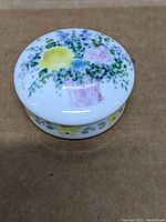 Top view of round trinket box lid showing pastel floral arrangement with yellow, blue, pink flowers and green leaves.
