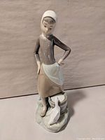 Front view of Lladro porcelain figurine showing girl with hands on hips, wearing brown dress and white apron and cap, standing next to milk bucket and white duck.