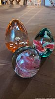 Three signed glass art paperweights displayed on a fabric surface. Tall peach egg-shaped, green/rose egg-shaped with flower, and round pink swirl design.