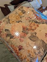 Floral patterned comforter folded and packaged in clear plastic showing muted orange, beige, brown, and green flower and leaf design.