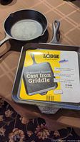 Two Lodge cast iron pans on brown tablecloth: an 11-inch seasoned square griddle with label and an 8-inch round frying pan underneath.