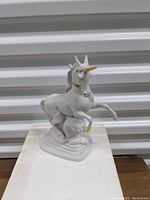 Front view of white unicorn figurine with gold horn and collar on plain surface with white ridged background.