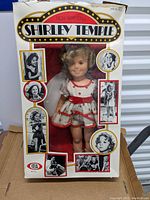 Front view of the Shirley Temple doll in its original box showing the doll dressed in a red and white dress with curls and surrounded by black and white photos of Shirley Temple.