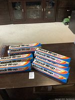 Photo showing 8 unopened boxes of Handy-Wrap plastic cling wrap arranged on a dark surface.