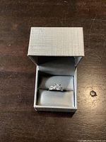 Silver textured jewelry box with the ring inside, showing the central clear gemstone and side stones from a top angle.