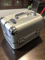 Closed metal makeup case showing silver ridged metal panels, metal corner protectors, chrome handle, and locking latches.