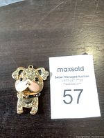 Front view of the gold-tone puppy dog charm featuring clear rhinestones and black rhinestone eyes and nose placed on a wooden surface next to a MaxSold auction card.