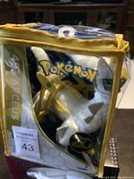 Photo showing the Arceus plush inside a clear zipper bag protector with yellow trim, front view.