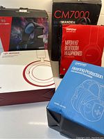 Five headphone boxes grouped on a table showing brand names and images of headphones including Commander CM7000, MPOW H7, MPOW Hearing Protection, Hunterspider Boltune, and MPOW Bluetooth headphones.
