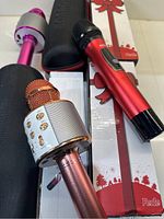 Three karaoke microphones arranged on a white surface with soft black carrying case and two holiday-themed packaging boxes. Details of microphones and accessories visible.