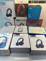 Photo showing boxed kids headphones from brands iClever, MPOW, B39, stacked together, various models and colors, mainly wireless and wired kids headphones.